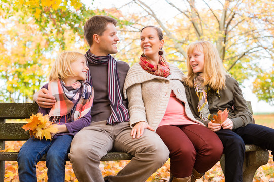 快乐的家庭，母亲，父亲和女儿或孩子坐在户外的长凳上，五颜六色的叶子和树下在秋天或秋天