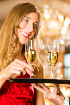 在一家高级餐厅，服务员端上香槟酒杯放在托盘上，一位女士端上一个杯子，背景是一盏大吊灯