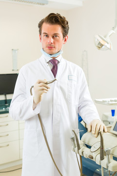牙医在他的手术中拿着一个钻头，看着观众，背景是牙医的工具