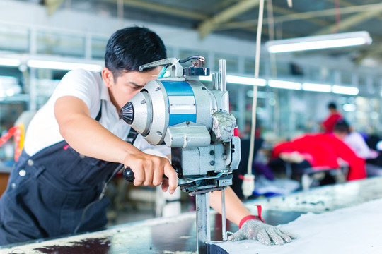 在一家亚洲纺织厂，一名印度尼西亚工人正在使用切割机（一种用于切割织物的大型机器），他戴着链子手套