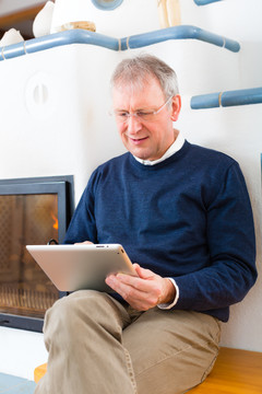 生活质量-老人或退休人员坐在炉子前的家里，在平板电脑上写电子邮件或读电子书