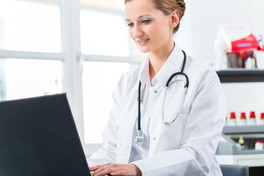 年轻的女医生坐在诊所窗口前的书桌旁用电脑或笔记本电脑写字或看书