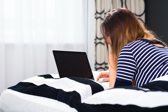 年轻女子躺在酒店房间的床上，她正在度假，用房间里的wifi与电脑上网
