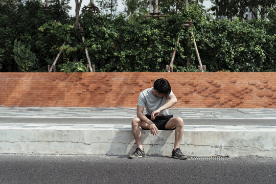 运动后在人行道上休息的疲倦的人。亚洲年轻人用衬衫擦脸。跑步后休息。