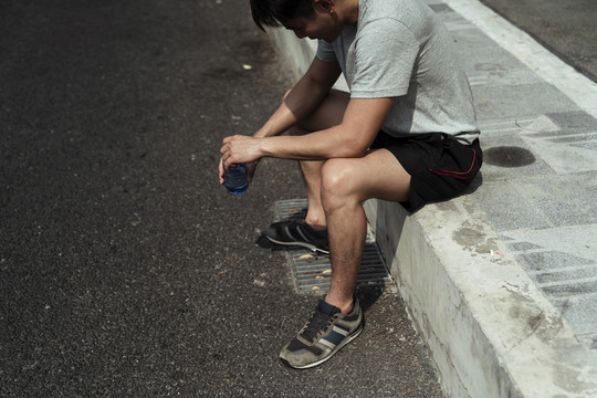 疲惫的运动员男子坐在人行道上，手里拿着一瓶水，看着地面。