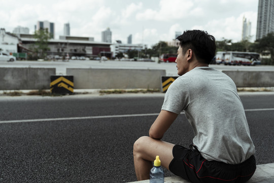 一个亚洲年轻人坐在路边，旁边拿着一瓶水，看着路。
