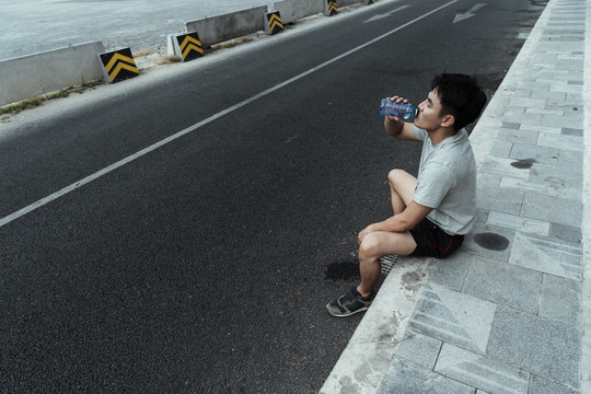 在赛道上跑步后坐在人行道上喝着一瓶水的疲惫的家伙。