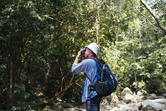 戴着帽子的男性徒步旅行者在森林里的岩石上散步，并为鸟类和树木拍照。