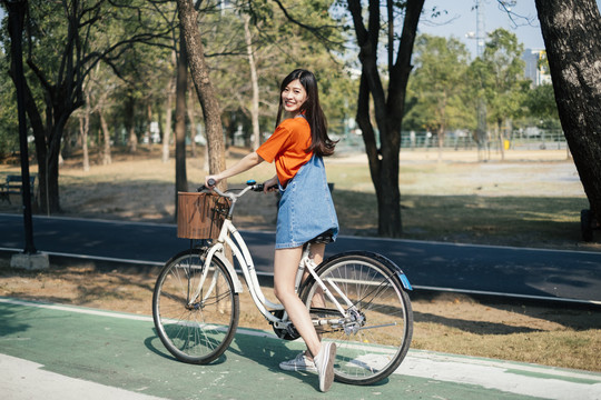 长发女孩穿着橙色t恤和牛仔裤，在公园的自行车道上停下自行车，对着镜头微笑。