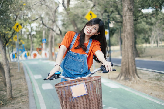 长发女孩穿着橙色t恤和牛仔裤在公园的自行车道上骑自行车，靠近镜头。