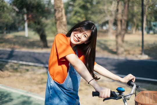 长发女孩穿着橙色t恤和牛仔裤，在公园的自行车道上停下自行车，然后歪着头看着镜头，微笑着。