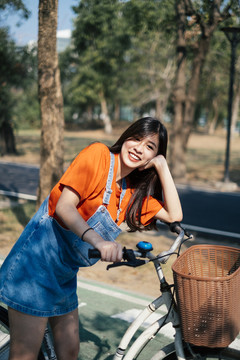 一个长发女孩穿着橙色t恤和牛仔裤，在公园的自行车道上停下她的自行车，然后把她的头放在自行车上。