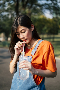身着橘色t恤和牛仔裤套头衫的长发女孩站在公园的阳光下，喝着水草的肖像。