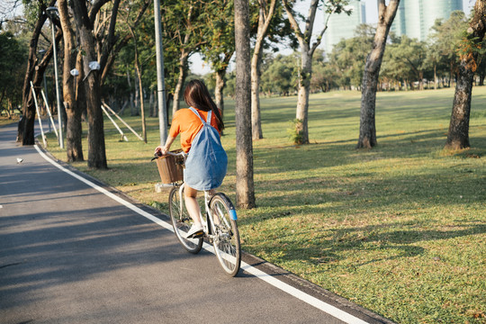 一个穿着橙色t恤和牛仔裤的长发女孩骑着自行车在公园的马路上。