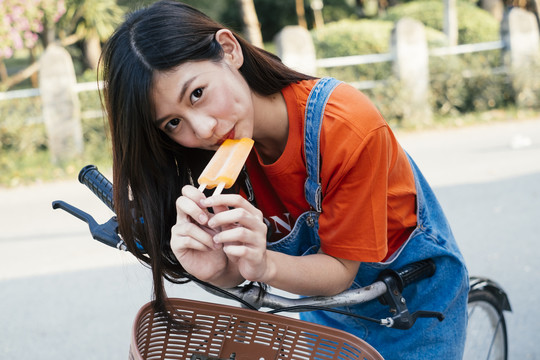 长发女孩穿着橘色t恤和牛仔裤在自行车上吃橘色冰淇淋，横。