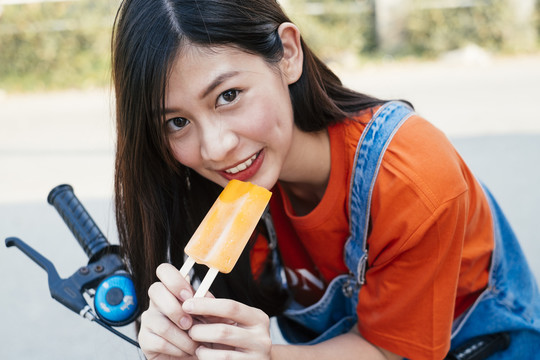穿着橙色t恤和牛仔裤套头衫的长发女孩拿着冰淇淋坐在自行车上。