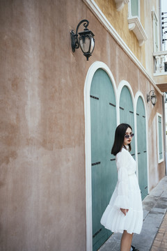 美丽的白衣长发女孩沿着绿色窗户和台灯的棕色建筑走着，回头看镜头。