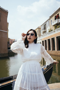 美丽的白衣长发女孩站在河边弯腰向后靠在木船上，抓起墨镜。