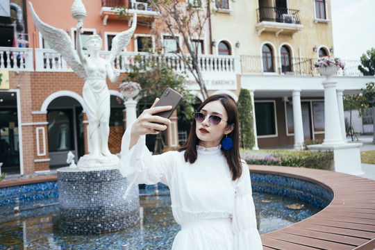 美丽的白衣长发女孩在浪漫小镇的白色喷泉与天使用智能手机自拍。