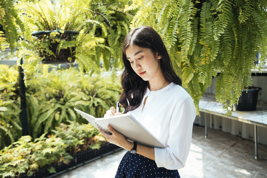 美丽的亚洲女人在热带花园的笔记本上写字。
