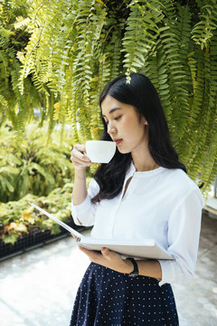 年轻的亚洲女人在花园里喝着一杯咖啡，读着一本书。