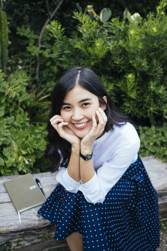 一幅年轻的亚洲女人的肖像，穿着白裙子和蓝裙子，坐在木凳上，两手托着脸，微笑着。