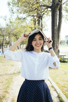 嬉皮的亚洲年轻女子，穿着白衬衫，微笑着把书放在头上。