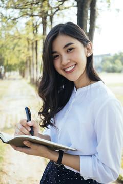 快乐的亚洲女人穿着白衬衫写着一本书，带着快乐的感觉，笑着笑着。小说作家微笑着写作的女孩。