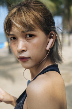 在阳光明媚、多风的日子里，戴着耳机的运动型短发女子在椰子树下的海滩上锻炼的特写镜头。