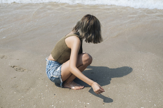 穿着短牛仔裤的短发女孩坐在海边的海滩上收集贝壳。