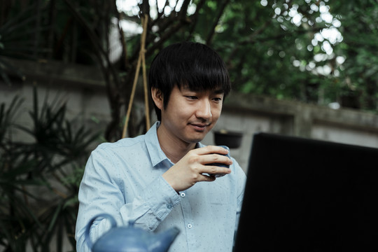 穿蓝衬衫的黑发男子拿着笔记本电脑在花园里工作，喝着茶。