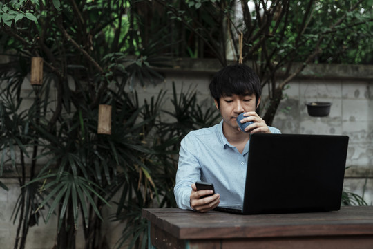 黑发男子穿着蓝色衬衫坐在花园里，看着手机查看顾客的订单，喝着热咖啡。