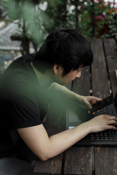 以绿叶为前景，拍摄了一张身穿黑色t恤的男子在工作时注视智能手机的高角度照片。