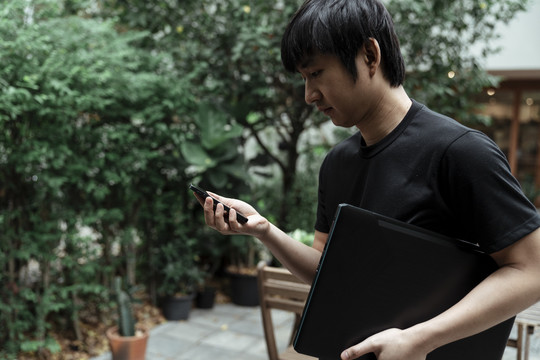 一名身穿黑色t恤的男子看着右手上的智能手机，左手拿着笔记本电脑。