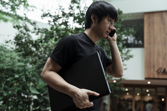 一名身穿黑色t恤的男子左手拿着笔记本电脑，一边和朋友通电话。
