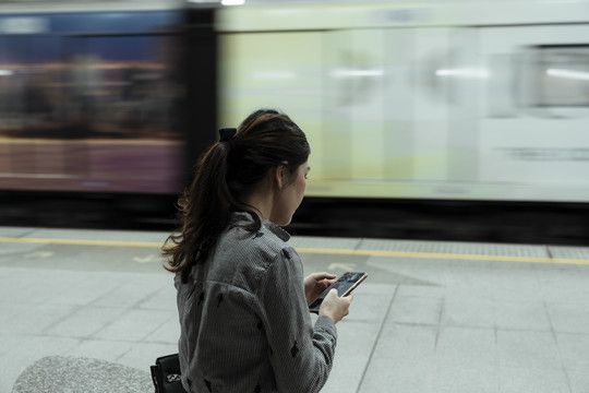 一名亚泰妇女坐在火车站台的长凳上，用智能手机等待火车回家。