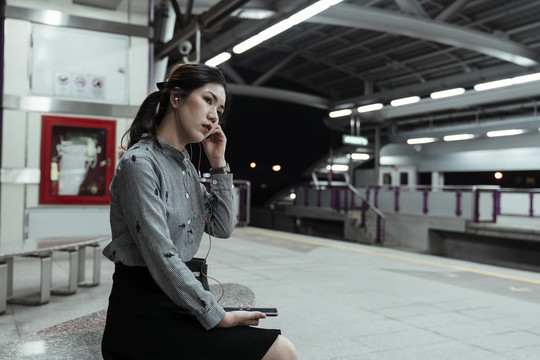 泰国亚裔妇女在曼谷的月台上等火车，晚上用耳机听智能手机里的音乐。