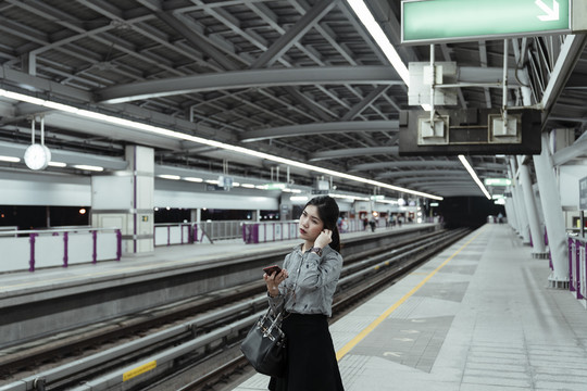 晚上，亚泰妇女在火车站台上用耳机听智能手机播放的音乐。