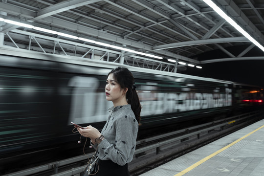 亚泰女子手持智能手机站在火车站台上，晚上等火车回家。
