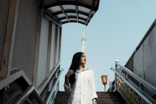 阳光下站在桥的楼梯上的一位身穿白衬衫的矮角黑发女子。