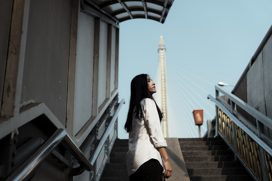 矮角的黑长发白衣女子站在阳光下的桥楼梯上，仰望天空。