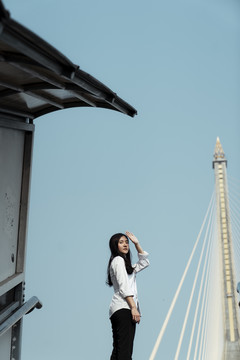 黑发白衣女子站在阳光下的桥上楼梯顶上，举手捂着眼睛。