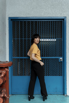 时尚写真-年轻的亚泰女性深色短发穿橙色衬衫过蓝色大门。后视图。侧视图。