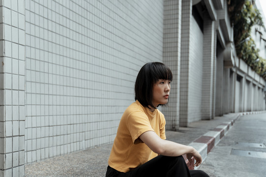 年轻的泰国亚裔女子深色短发，身穿橙色衬衫，坐在街上等待有人来接她。