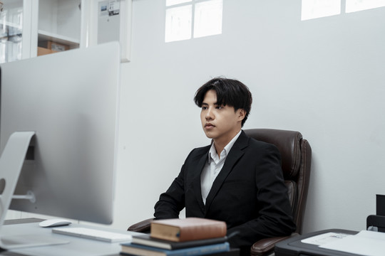 年轻的亚洲企业老板坐在椅子上看着办公室里的电脑。
