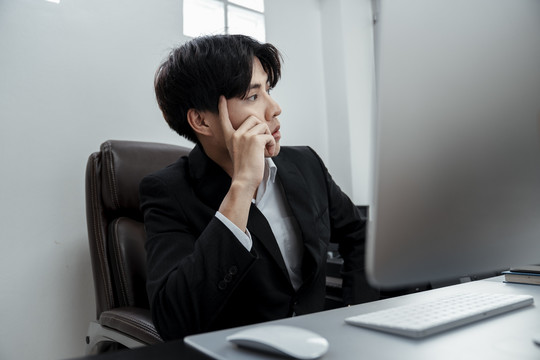 体贴的亚洲商人穿着黑色西装坐在电脑前的办公室椅子上工作。