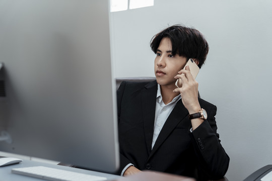 穿着黑色西装的亚洲帅哥在办公室办公桌上打电话。