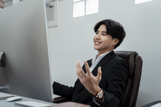 快乐的亚洲商人穿着黑色西装坐在椅子上看电脑。成功的商人对结果很满意。