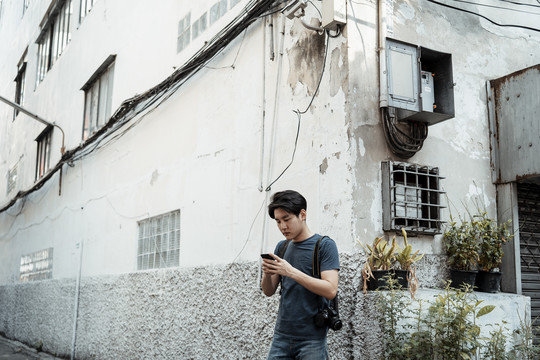 年轻英俊的亚洲摄影师在曼谷街头小巷使用智能手机。