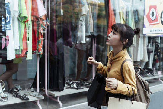 可爱的亚洲女人穿着黄色毛衣在街上的购物窗口。看着镜子后面挂着的衣服。购物窗口。